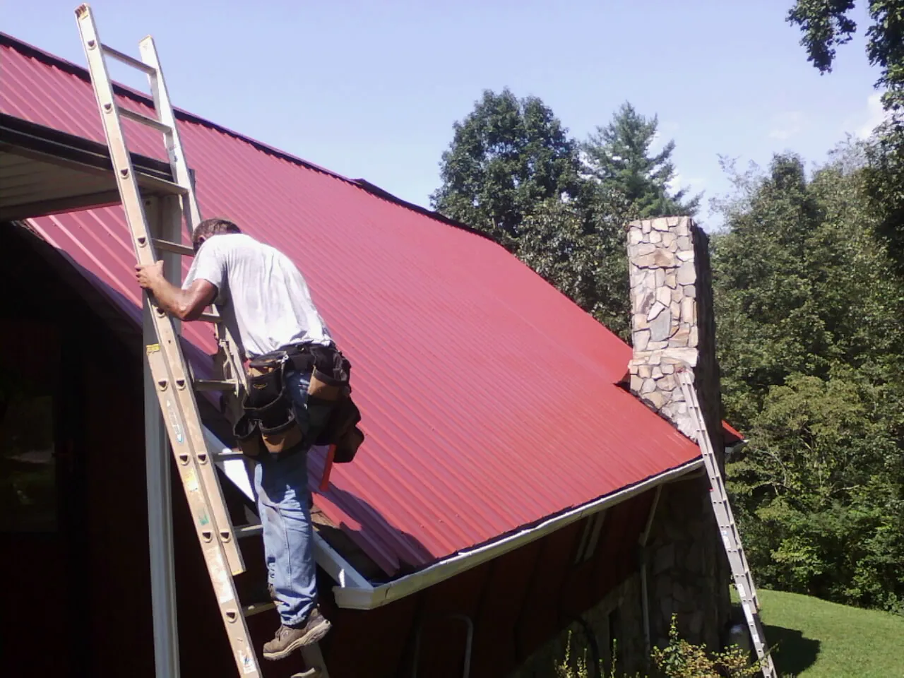 Roofer on a ladder beside a red metal roof and stone chimney — Soffit & Fascia Repair by Roofing Brothers in Millwood